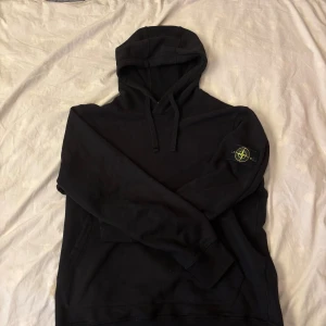 Svart hoodie från Stone Island  - Svart hoodie från Stone Island i storlek XL med klassisk patch på vänster arm. Tröjan har en stor och bekväm luva med snörning och känguruficka fram. Mjuk insida och stilren design, perfekt för alla stilar!                                                                                       Tar emot rimliga byten! Tveka inte på att fråga om tröjan, kan skicka fler bilder, mått osv. Säljer pga att jag har liknande tröjor som används mer. Skickar snabbt!