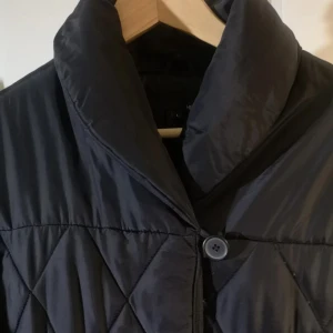 Svart quiltad pufferjacka Från H&M Stl L - Svart pufferjacka från Hennes i storlek L med snyggt quiltat mönster och bred krage. Jackan har knäppning med stora knappar framtill och är tillverkad i syntetmaterial. Perfekt för dig som gillar stilren och enkel design.