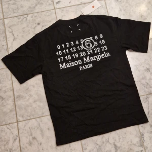 Maison Margiela svart t-shirt med tryck - Svart t-shirt från Maison Margiela med ikoniskt siffror-tryck och logga på ryggen. Klassisk rund hals, korta ärmar och diskreta vita stygn på bröstet. Tillverkad i mjuk bomull för en skön känsla. Storlek M. Perfekt för dig som gillar streetstyle och designerplagg. Tryck på ryggen och den ikoniska stitch loggan på framsidan. Hör av er vid minsta fråga 🤍🙌