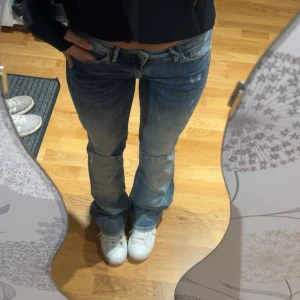 Blå bootcut jeans med slitningar - Säljer ett par blå bootcut jeans med låg midja och slitna detaljer vid knäna. Jeansen har klassisk femficksdesign och är tillverkade i jeansmaterial. De har en snygg tvätt med ljusare partier och råa kanter vid hålen för en cool look. Midje: 34-35cm och innebenslängd: 75-77cm!!