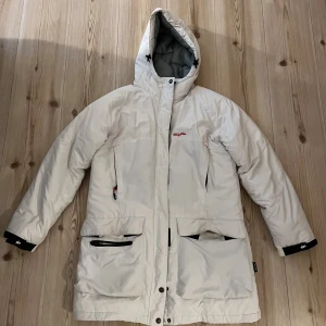 Vit täckjacka från Windy Peak - Säljer en vit dunjacka från Windy Peak med huva och flera fickor med dragkedja. Jackan har quiltad insida, lång ärm och snygga svarta detaljer vid ärmslut. Perfekt för kalla dagar och har en clean, stilren look.