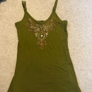 topp med pärlor - Snygg olivgrön linnetopp med smala axelband och v-ringad hals. Framsidan har detaljerad broderi med guldiga pärlor och paljetter i ett unikt mönster. Toppen är lätt och mjuk, perfekt för varma dagar. Materialet känns som bomull och toppen har en figurnära passform.