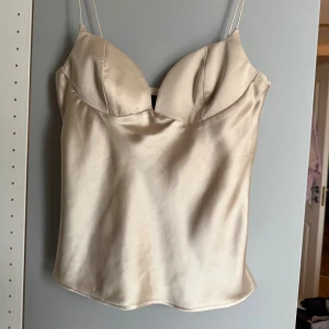 Champagnefärgad satin korsett-topp - Snygg korsett-topp i champagnefärgad satin med tunna axelband och formade kupor. Helt ny och aldrig använd!