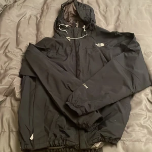 Svart vindjacka från The North Face XL - Svart vindjacka från The North Face i storlek XL. Jackan har huva med snörning, broderad logga på bröstet och elastiska muddar. Materialet är nylon och polyester, vilket gör den lätt och smidig. Perfekt för dig som gillar stilren och funktionell design.