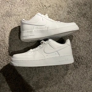 Nike Air Force 1 helvita sneakers - Klassiska Nike Air Force 1 sneakers i helvitt skinn med lågt skaft. Snygg och clean design med perforerad tå, vit sula och ikoniska swoosh-loggan på sidorna. Perfekta för dig som vill ha en tidlös och fräsch look.