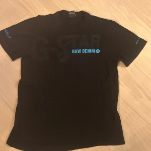Svart G-Star t-shirt med blå text - Svart t-shirt från G-Star i storlek S med coolt tryck 'RAW DENIM' i blått på bröstet och G-Star-logga på ärmen. Klassisk rund hals och korta ärmar. Skön bomullskvalitet som passar perfekt till jeans eller shorts.