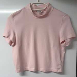 Fin Ljusrosa polotopp från Monki - Säljer en ljusrosa polotopp från Monki i storlek S. Toppen är croppad med korta ärmar och har en mjuk polokrage. Tillverkad i stretchig bomull med 5% elastan för extra komfort. Perfekt för dig som gillar enkel och clean stil.