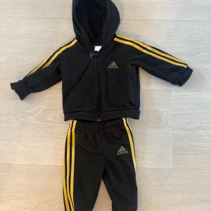 Adidasdress bebis 0-3 mån guld och svart  - Svarta mjukisbyxor från Adidas med klassiska gula ränder längs sidorna och Adidas-logga på benet. 