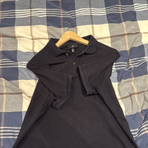 Svart pikétröja från H&M - Svart pikétröja från H&M med klassisk krage och två knappar framtill. Tröjan är tillverkad i bomull och har en diskret ribbad struktur som ger en snygg och stilren look. Perfekt för dig som gillar enkel och clean stil.