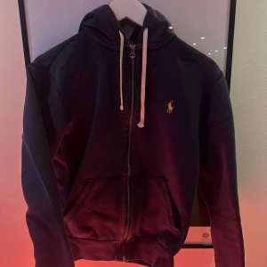  Ralph Lauren - Snygg marinblå hoodie från Polo Ralph Lauren med gul broderad logga på bröstet. Tröjan har huva med vita snören, dragkedja framtill och två fickor. Perfekt för chill dagar och har en klassisk streetstil. Kan vara lite ljusare på vissa ställen och har lite skav på metallen :) 