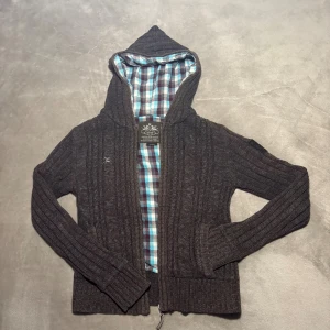 Stickad Cardigan - Mysig brun stickad hoodie med dragkedja och huva. Insidan av huvan är fodrad med rutigt tyg i blått, vitt och brunt. Tröjan har långa ärmar och ribbade muddar, perfekt för kyliga dagar. Den har ett hål på insidan vid armhålan som man ser på bild 4. Står S men fråga innan om mått om du är intresserad.🦦