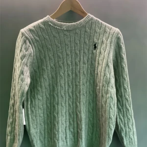 Grå kabelstickad tröja Polo Ralph Lauren - Kabelstickad grå tröja från Polo Ralph Lauren med klassisk rund halsringning och broderad logga på bröstet. Tillverkad i 100% bomull och har långa ärmar. Perfekt för dig som gillar en stilren och tidlös look.