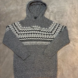 Grå stickad hoodie med mönster - Mysig grå stickad hoodie med vit och svart zigzag-mönster över bröst och ärmar. Tröjan har huva med snörning och långa ärmar. Perfekt för kalla dagar när du vill vara både varm och snygg.