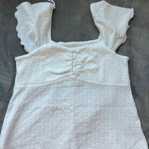 Vit blus från Feather & Bone - Supersöt vit blus från Feather & Bone med volangärmar och smockad struktur. Blusen har dekorativa knappar framtill och fyrkantig ringning. Perfekt för sommarens alla tillfällen och ger en fräsch och trendig look.