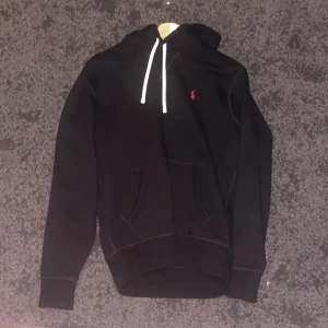 Svart hoodie från Polo Ralph Lauren - Svart hoodie från Polo Ralph Lauren med vit snörning och röd logga på bröstet. Tröjan har huva, känguruficka och långa ärmar. Perfekt för chill dagar och enkel att matcha med jeans eller joggers. Använd endast 1 gång och köpt från Zalando.