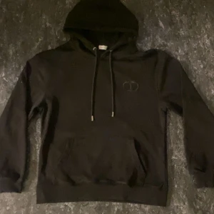 Svart CD hoodie med broderad logga - Svart hoodie med broderad CD-logga på bröstet. Tröjan har huva med snörning, magficka och ribbade muddar vid ärmslut och nederkant. Perfekt för dig som gillar stilrena och exklusiva streetwear-plagg.