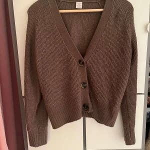 Brun stickad kofta från Lindex - Mysig brun stickad kofta från Lindex med grov ribbad struktur och tre stora knappar framtill. Koftan har v-ringad hals och långa ärmar, perfekt för lager-på-lager under kyliga dagar. Enkel och stilren design som är lätt att matcha.