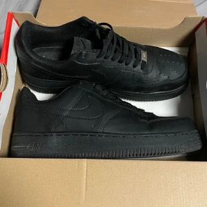 Svarta Nike Air Force 1 sneakers - Säljer ett par helsvarta Nike Air Force 1 sneakers med klassisk låg siluett. Skorna har skinnöverdel, perforerad tå och svart sula. Perfekta för en clean och stilren look. Ikonisk Nike-logga på sidan och snörning framtill.