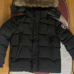 Canada Goose Wyndham  - Säljer en svart dunjacka från Canada Goose med pälskantad huva och klassisk patch på ärmen. Jackan är quiltad, har dragkedja och knappar framtill samt flera praktiska fickor. Perfekt för kalla vinterdagar och riktigt snygg streetstil.