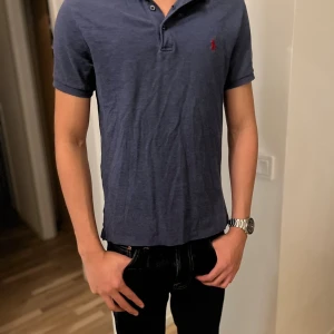 Blå slim fit piké från Polo Ralph Lauren - Snygg mörkblå pikétröja från Polo Ralph Lauren i slim fit med stretch mesh-material. Klassisk krage, korta ärmar och knappar framtill. Röd broderad logga på bröstet ger en clean och stilren look. Perfekt för dig som gillar enkel och tidlös stil.