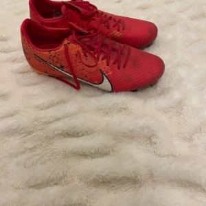 Nike Mercurial Air Zoom fotbollsskor röda - Säljer ett par Nike Mercurial Air Zoom fotbollsskor i rött och orange med svarta och vita detaljer. Skorna har snörning, platt sula med dobbar och är tillverkade i syntetmaterial. Snyggt grafiskt mönster på sidorna och klassisk Nike-logga.