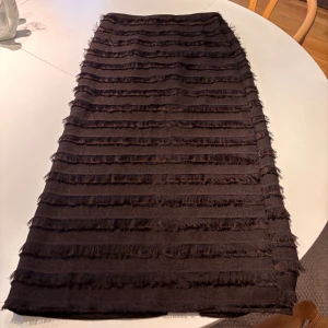 Brun långkjol med fransar från H&M Edition - Säljer en brun långkjol från H&M Edition i storlek 34. Kjolen har horisontella fransade ränder och en slits bak för extra rörelsefrihet. Materialet känns mjukt och har en cool struktur som sticker ut. Perfekt för dig som gillar unika detaljer och vill ha något annorlunda i garderoben.