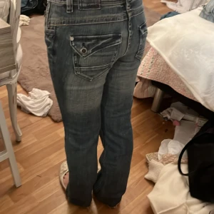 Blå bootcut jeans med coola fickor - Säljer ett par blå jeans med bootcut passform och snygga detaljerade bakfickor med lock och knapp. Jeansen har låg midja och slitna kanter vid benslut för en avslappnad vibe. Materialet är klassisk denim och färgen är tvättad blå med ljusare partier.