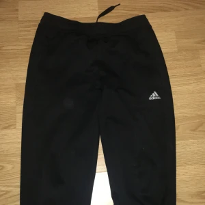 Svarta mjukisbyxor från Adidas - Svarta mjukisbyxor från Adidas med klassisk logga på benet. Byxorna har snörning i midjan och är gjorda i mjukt syntetmaterial, perfekta för chill eller träning. Snygg, enkel design med smal passform och diskret tryck vid benslutet.