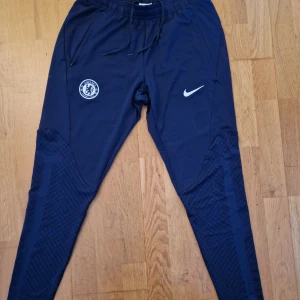 Chelsea Nike Dri-Fit träningsbyxor  - Mörkblå Chelsea FC träningsbyxor från Nike med Dri-Fit-teknologi. Slim fit-modell med snörning i midjan. Snyggt tryck med klubbmärke och Nike-logga. Använd fåtal gånger så I princip väldigt nya!