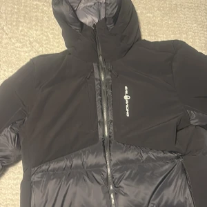Svart pufferjacka från Sail Racing - Säljer en svart pufferjacka från Sail Racing i storlek Large. Jackan har huva, flera dragkedjor och en stilren design med Sail Racing-logga på bröstet. Materialet är syntet och polyester, perfekt för kalla dagar. Jackan har lång ärm och quiltade partier för extra värme.