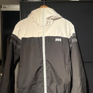 Svart och vit skidjacka från Helly Hansen - Snygg vindjacka från Helly Hansen i svart och vitt med huva och hel dragkedja. Jackan har RECCO-reflektor på ärmen, meshfoder och coola detaljer på axlarna. Perfekt för dig som gillar stilren och sportig look.