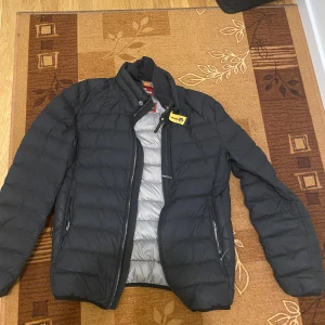 Svart dunjacka Parajumpers - Svart dunjacka från Parajumpers med quiltad design och gul detalj vid kragen. Jackan har dragkedja framtill och två sidofickor med dragkedja. Insidan är ljusgrå och jackan har en hög krage som ger extra skydd mot vind. ( PRIS KAN DISKUTERAS ) 