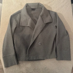 Grå kort rock med bred krage - Snygg grå kort rock med bred krage och flera knappar framtill. Jackan har en stilren och minimalistisk design, med raka linjer och lite boxig passform. Perfekt för dig som gillar klassisk stil med modern twist. Materialet känns som ull och håller dig varm under kyliga dagar.