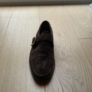 Bruna mocka skor från Loake - Snygga bruna loafers från Loake i mjuk mocka med klassiskt spänne i guld. Skorna har en elegant och tidlös design med rund tå och platt sula, perfekta för dig som gillar stilrena detaljer och hög kvalitet.