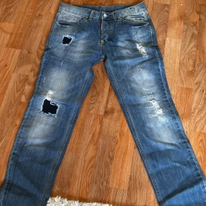Blå Dsquared jeans med slitningar - Säljer ett par blå jeans från Dsquared med coola slitningar och hål på låret och knät. Jeansen har klassisk femficksdesign, raka ben och snyggt tvättad finish. Perfekta för dig som gillar en avslappnad och trendig look.