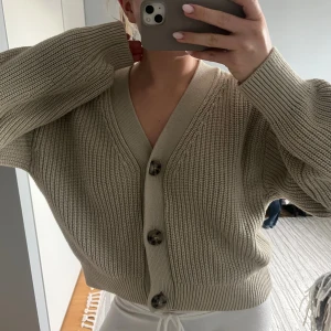 Beige stickad kofta med knappar - Säljer en beige stickad kofta från HM med grov ribbad struktur och stora mörka knappar framtill. Koftan har v-ringad hals och långa ärmar med breda muddar. Perfekt för lager-på-lager och chill stil.