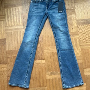 True Religion Becca Bootcut Jeans W26 - Oanvända blå jeans från True Religion, modell Becca, midrise bootcut flap. Prislappar finns fortfarande kvar. Jeansen har klassisk femficksdesign, coola bakfickor med lock och gul kontrastsöm samt silverfärgade knappar. Tillverkade i jeansmaterial med en lätt tvättad look och markerade sömmar. Gula och vita sömmar.