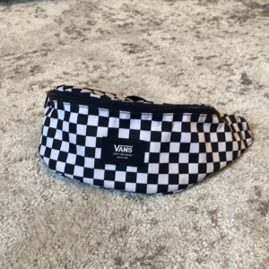 Svartvit rutig axelväska från Vans - Säljer en schysst axelväska från Vans med klassiskt svartvitt rutigt mönster. Väskan har två olika fack. Ett lite mindre som passar till telefonen och ett stort för till exempel nycklar, plånbok och annat. Väskan har justerbar rem så du kan bära den både över axeln eller runt midjan. 