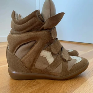 Beige höga sneakers med kardborre - Säljer ett par höga sneakers i beige och vitt med coola paneler och flera kardborreband framtill. Skorna har en plattformssula och är tillverkade i mocka och skinn, vilket ger dem en unik och trendig look. Perfekta för dig som gillar statement-skor med extra höjd.