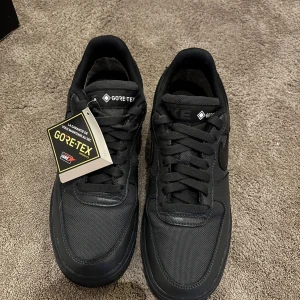 Nike Air Force 1 Gore-Tex  - Svarta Nike Air Force 1 med Gore-Tex-material som gör dem vattentäta. Swoosh-logga på sidan och Gore-Tex-brodyr på hälen. Perfekta för regniga dagar.