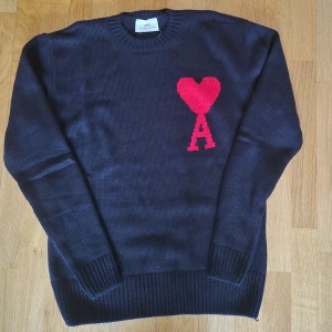 Svart stickad tröja från Ami Paris - Svart stickad tröja från Ami Paris med röd hjärt- och A-detalj på bröstet. Tröjan är i 100% bomull, har rund hals och långa ärmar. Perfekt för dig som gillar stilrena plagg med en twist. Ribbstickade muddar vid ärmslut och nederkant.