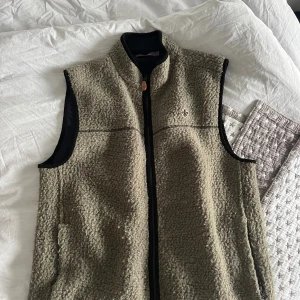 Teddy vest Morris  - Säljer en kahki fleeceväst från Morris med hög krage och hel dragkedja framtill. Västen har en mjuk, teddy-liknande yta och svarta detaljer vid ärmöppningarna. Perfekt för lager-på-lager och snygg med broderad logga på bröstet.