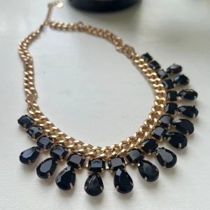 Guldigt halsband med svarta stenar - Statement-halsband med tjock guldfärgad kedja och droppformade svarta stenar som hänger längs hela kedjan. Perfekt för att lyfta din outfit med en cool och glam touch. #halsband #accessoarer