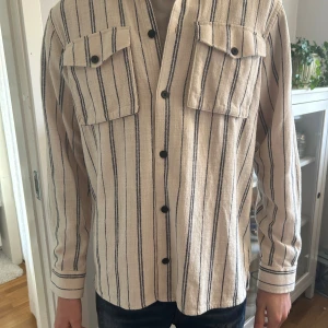 Randig overshirt från Casual Friday - Snygg beige overshirt från Casual Friday i storlek M med svarta vertikala ränder. Skjortan har två bröstfickor med knappar, klassisk krage och långa ärmar med knappslå. Materialet är mjukt och känns som bomull, perfekt för en chill och stilren look. Använd fåtal gånger så overshirten är i topp skick.