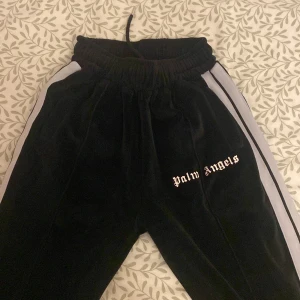 Svarta Palm Angels mjukisbyxor med vita ränder och dragkedja vid längden  - Svarta mjukisbyxor från Palm Angels med vita sidoränder och broderad logga på benet. Byxorna har resår i midjan med snörning och är tillverkade i ett mjukt, sammets material. Sitter som s/m 
