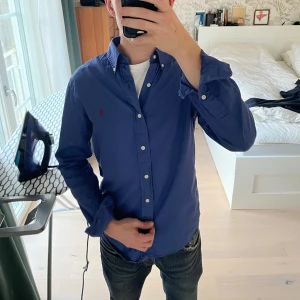 Blå skjorta från Ralph Lauren - Klassisk blå skjorta från Ralph Lauren med röd broderad logga på bröstet. Skjortan har lång ärm, knappar hela vägen och button-down krage. Tillverkad i mjuk bomull och har en slim passform som sitter snyggt på kroppen.