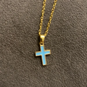 Guldigt halsband med blått kors - Snyggt halsband i guldtonad rostfritt stål med ett korsformat hänge i ljusblå färg. Kedjan är tunn och har en klassisk karbinlås. Perfekt för dig som gillar stilrena smycken med en färgklick.