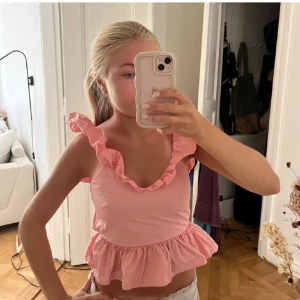 Rosa volangblus med öppen rygg - Supersöt rosa blus med volangdetaljer längs axelbanden och v-ringad hals. Toppen har öppen rygg med knytband och en peplumkant nedtill. Perfekt för dig som gillar romantisk och lekfull stil. Materialet känns lätt och luftigt.