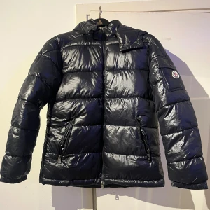 Svart pufferjacka från Moncler - Säljer en svart pufferjacka från Moncler med glansig finish och klassisk logga på ärmen. Jackan har två dragkedjeförsedda fickor, hög krage och quiltad design. Perfekt för kalla dagar och har en riktigt snygg streetstil.  Är själv 180 och den passar perfekt, helt oanvänd köpte den för 3 veckor sedan.