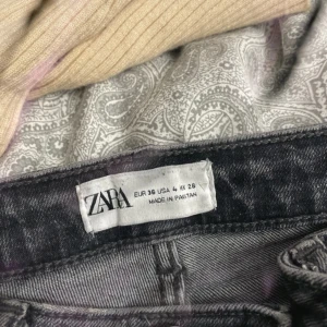 Grå jeans med slitningar från Zara - Säljer ett par grå jeans från Zara i klassisk femficksmodell. Jeansen har snygga slitningar och råa kanter vid knäna för en trendig look. Modellen är rak och midjehög, perfekt till sneakers eller boots. Materialet är mjukt denim i bomull.